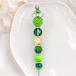 May include: A decorative pen featuring a vertical arrangement of beads in various shades of green. The design includes rhinestone-encrusted spheres, a floral-patterned bead, and a crown-shaped topper with a green gem, all separated by silver spacers.