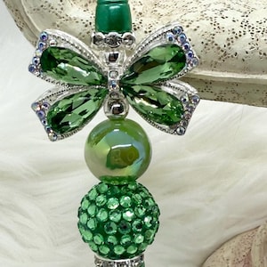 May include: A green bottle stopper with a butterfly design. The butterfly has green crystal wings and a silver frame with iridescent accents. The stopper has a green, iridescent bead and a green, gem-studded bead.