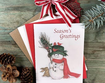 May include: Christmas cards featuring a snowman illustration, a red scarf, and the text "Season's Greetings." The cards are paired with a red and white striped bow, pine cones, and a sprig of evergreen.