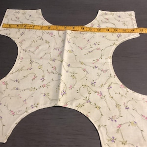 May include: A cream-colored, floral-patterned fabric piece with a tape measure laid across it. The fabric features a delicate design of vines and small flowers in pink, purple, and green. The tape measure shows a measurement of approximately 17 inches.