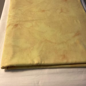 May include: Folded yellow fabric with a mottled pattern, possibly dyed or printed. The fabric has a soft, muted color palette with hints of brown and tan. The fabric is folded neatly on a white surface.