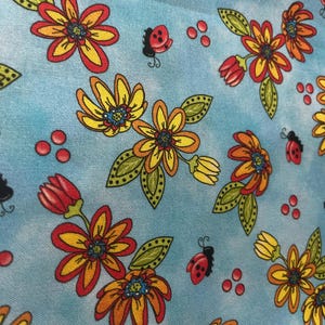 May include: A light blue fabric featuring a repeating pattern of yellow and orange flowers with green leaves, interspersed with small red ladybugs and clusters of three small red dots. The design has a soft, watercolor-like background texture.