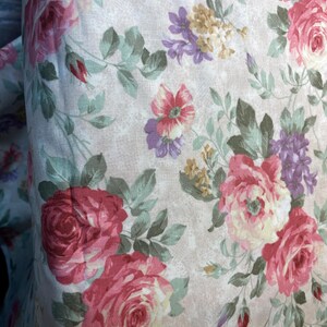 May include: A fabric featuring a floral pattern on a light beige background. The design includes large pink and cream roses, smaller purple flower clusters, and tiny yellow blossoms, all accented by sage green leaves and stems on a textured surface.