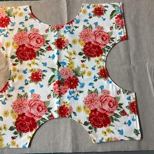 May include: A cut fabric piece with a floral pattern featuring red roses, pink dahlias, yellow daisies, and blue accents on a white background. The fabric is cut in a unique shape, possibly for a garment.