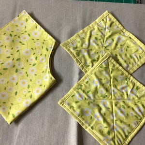 May include: Four yellow fabric items with a daisy floral pattern. One item is diamond-shaped with curved edges, and the other three are square with a yellow border. The daisy pattern includes white flowers and green leaves.