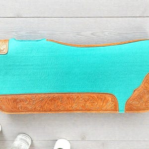May include: A turquoise saddle pad with brown leather accents. The leather features a floral tooled design on the corners and bottom edge. The pad is rectangular with rounded corners.