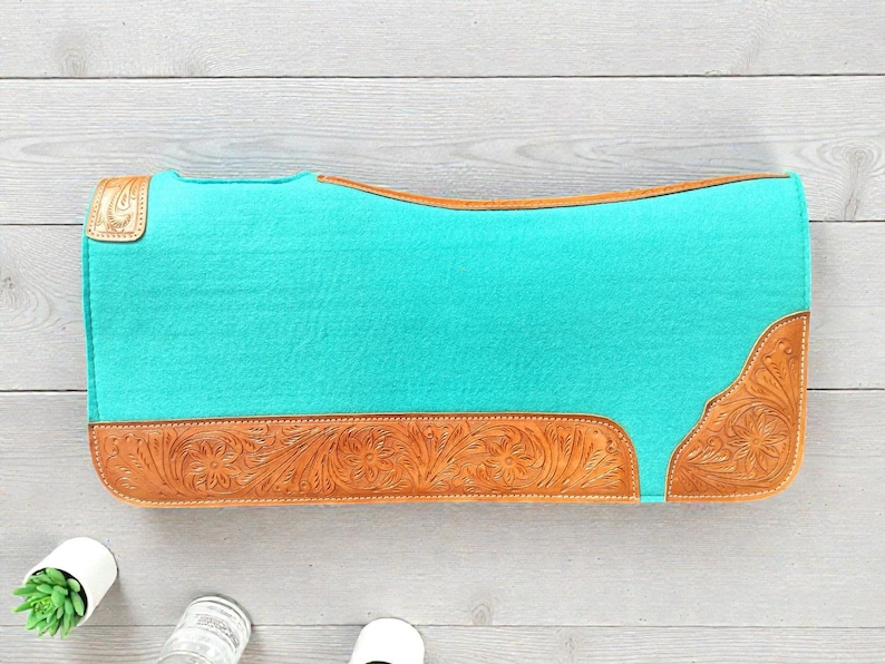 May include: A turquoise saddle pad with brown leather accents. The leather features a floral tooled design on the corners and bottom edge. The pad is rectangular with rounded corners.