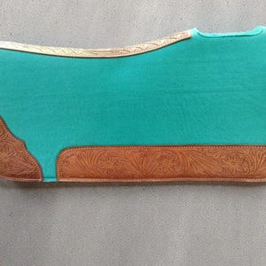 May include: A turquoise saddle pad with brown leather accents. The leather features floral and scroll tooling along the edges and corners. The pad is rectangular with a curved top edge.
