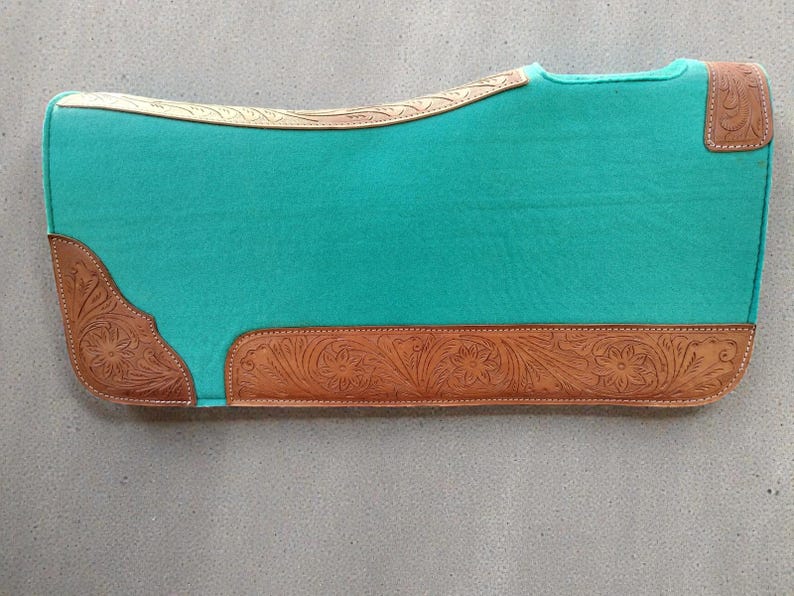 May include: A turquoise saddle pad with brown leather accents. The leather features floral and scroll tooling along the edges and corners. The pad is rectangular with a curved top edge.