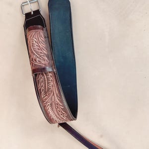 Puede incluir: Dos correas de cuero con hebillas metálicas. Una correa es de color marrón oscuro con un diseño floral tallado, la otra es negra. Las correas están conectadas por una correa más pequeña, de color marrón oscuro, con una hebilla.