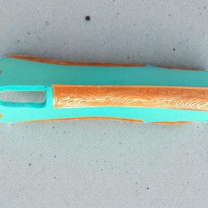 May include: A turquoise-colored leather item with a rectangular shape and a cutout. It features decorative, embossed brown leather accents with floral patterns on the edges and center. The item appears to be a protective cover or sleeve.
