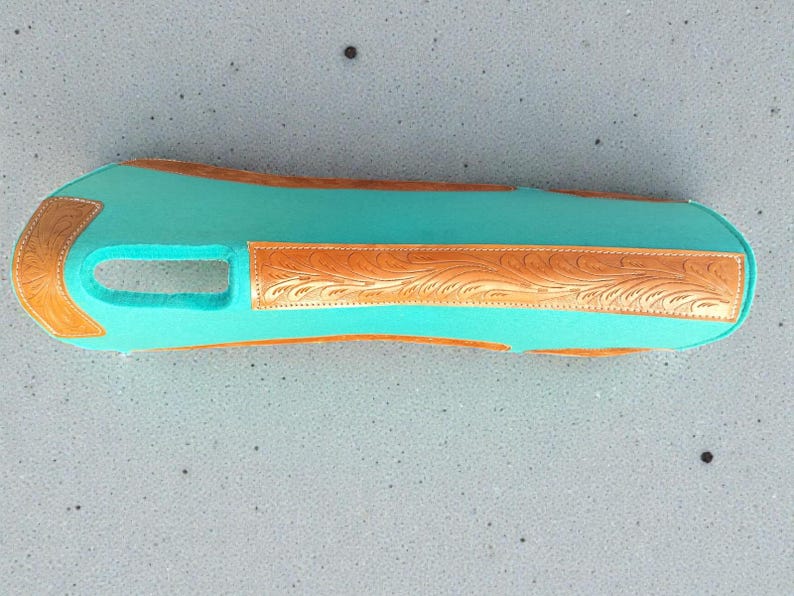 May include: A turquoise-colored leather item with a rectangular shape and a cutout. It features decorative, embossed brown leather accents with floral patterns on the edges and center. The item appears to be a protective cover or sleeve.