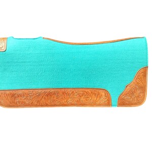 May include: A turquoise saddle pad with a rectangular shape. The pad features a light brown, tooled leather border with floral designs. The leather accents are on the bottom and corners of the pad.