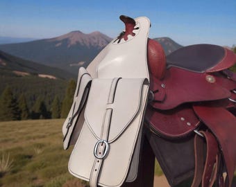 Puede incluir: Primer plano de una alforja de cuero blanco con costuras negras y hebilla plateada, sujeta a una silla de montar de cuero burdeos. La silla está montada sobre un poste de madera, con un fondo montañoso.