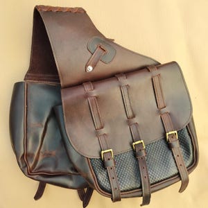 May include: A brown leather saddlebag with a flap and three buckle closures. The bag has a textured black panel and a shoulder strap. The leather is a rich, dark brown color, and the buckles are gold-colored.