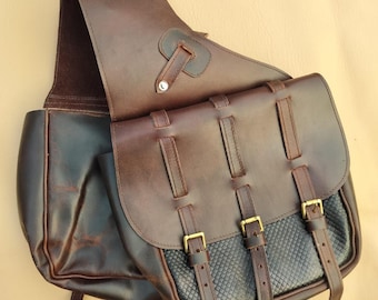 Puede incluir: Alforja de cuero marrón con solapa y tres cierres de hebilla. La bolsa tiene un panel negro texturizado y una correa para el hombro. El cuero es de un color marrón oscuro intenso y las hebillas son doradas.
