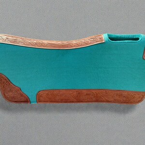 May include: A turquoise saddle pad with brown leather accents. The pad features a curved shape with a cutout for the saddle horn. The leather accents are embossed with a floral pattern. The pad is designed for equestrian use.