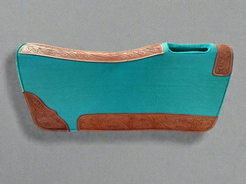 May include: A turquoise saddle pad with brown leather accents. The pad features a curved shape with a cutout for the saddle horn. The leather accents are embossed with a floral pattern. The pad is designed for equestrian use.