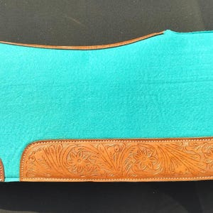 May include: A turquoise saddle pad with brown leather trim. The leather trim features a floral pattern. The saddle pad is rectangular with rounded corners and is designed for equestrian use.