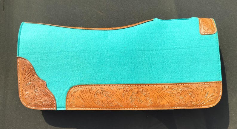 May include: A turquoise saddle pad with brown leather trim. The leather trim features a floral pattern. The saddle pad is rectangular with rounded corners and is designed for equestrian use.