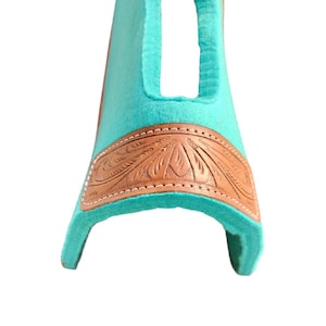 May include: A turquoise saddle pad with a brown leather accent featuring an embossed floral design. The leather is stitched to the turquoise pad with white thread. The pad has a curved shape with an opening at the top.
