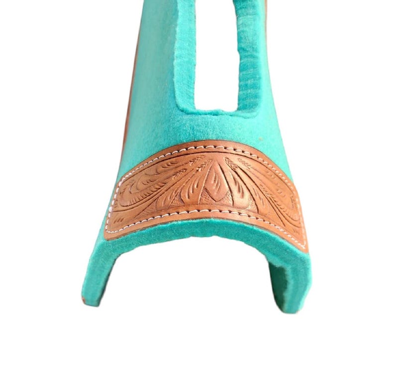 May include: A turquoise saddle pad with a brown leather accent featuring an embossed floral design. The leather is stitched to the turquoise pad with white thread. The pad has a curved shape with an opening at the top.