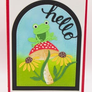 Tarjeta de saludo hecha a mano: Rana adorable sobre un hongo con flores, saludo con una escena natural encantadora, interior en blanco, tarjeta alegre y amigable.