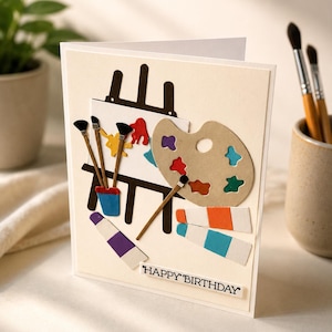 May include: A handmade birthday card with an artist theme. The card features a miniature easel with a painting, a palette, paint tubes, and brushes. The words "HAPPY BIRTHDAY" are printed on the bottom.