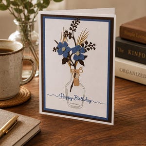 May include: A greeting card with a floral design. The card features blue flowers and brown stems arranged in a vase illustration. The words "Happy Birthday" are written in a script font. The card has a brown and blue border.