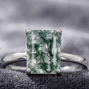 May include: A ring featuring a rectangular emerald-cut moss agate gemstone with dark green, fern-like inclusions set in a polished silver-toned metal band with four prongs securing the stone at each corner of the setting.