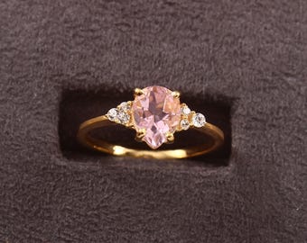 May include: A gold ring featuring a pear-shaped, light pink gemstone as the centerpiece. The ring is accented with small, round, clear gemstones on either side of the main stone. The ring is set against a dark gray background.