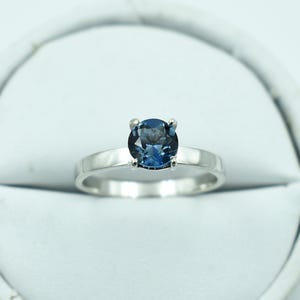 以下が含まれることがあります： 丸い、濃い青色の宝石をあしらったシルバーリング。 宝石は4つの爪でセットされており、バンドはシンプルな磨かれたシルバーです。 リングは白い背景に表示されています。