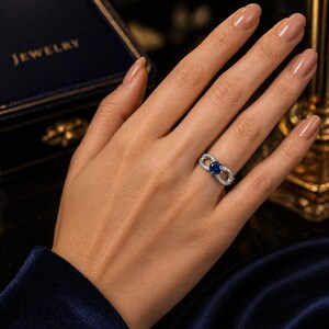 May include: A silver ring with a blue sapphire center stone and surrounding white gemstones. The ring is on a person's hand, with a dark blue velvet background and a jewelry box in the background.