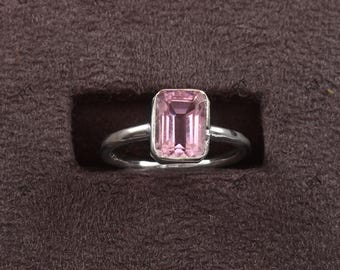 May include: A silver ring featuring a rectangular, light pink gemstone. The ring is set in a simple, elegant design, showcasing the clarity and color of the stone. The ring is displayed on a dark brown surface.