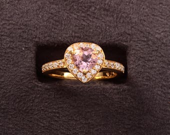 May include: A gold ring with a heart-shaped pink gemstone center, surrounded by a halo of small white gemstones. The band is also adorned with small white gemstones. The ring is set against a dark background.