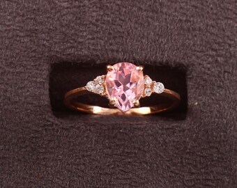 May include: A rose gold ring featuring a pear-shaped, light pink gemstone as the centerpiece. The ring is accented with smaller, round, clear gemstones on either side of the main stone. The ring is set against a dark brown background.