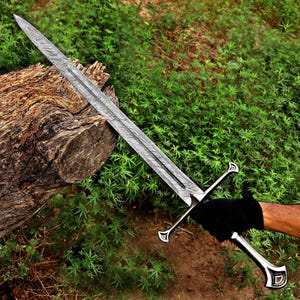 May include: An intricately designed sword with a patterned blade and a silver hilt. The sword is set against a backdrop of green foliage and a weathered log. The hilt includes a crossguard and a decorative pommel.