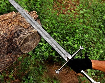 May include: A long, ornate sword with a patterned blade and a silver hilt. The sword is held against a backdrop of green foliage and a weathered log. The hilt features a crossguard and a decorative pommel.