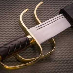 K&ouml;nnte beinhalten: Nahaufnahme eines Wing Chun Butterfly-Schwertes. Das Schwert hat eine polierte Stahlklinge, einen schwarzen Griff und einen goldfarbenen Parier. Das Schwert ist teilweise in einer schwarzen Scheide verstaut. Der Hintergrund ist eine strukturierte, dunkelgraue Oberfl&auml;che.