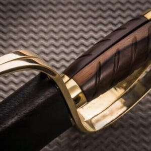 K&ouml;nnte beinhalten: Nahaufnahme eines Schwertgriffs. Der Griff ist aus dunkelbraunem Holz mit geschnitzten Rillen, umrahmt von goldfarbenem Metall. Die Klinge ist in schwarzes Leder geh&uuml;llt. Das Design deutet auf ein historisches oder Fantasy-Thema hin.