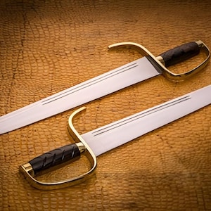 K&ouml;nnte beinhalten: Zwei Schwerter mit silbernen Klingen und Messinggriffen mit schwarzen Griffen liegen auf einer strukturierten, braunen Oberfl&auml;che. Die Klingen haben ein Doppelrillen-Design. Die Griffe haben einen gebogenen Handschutz.