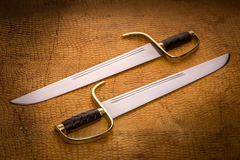 K&ouml;nnte beinhalten: Zwei Schwerter mit silbernen Klingen und Messinggriffen mit schwarzen Griffen liegen auf einer strukturierten, braunen Oberfl&auml;che. Die Klingen haben ein Doppelrillen-Design. Die Griffe haben einen gebogenen Handschutz.