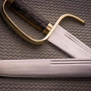 K&ouml;nnte beinhalten: Nahaufnahme eines S&auml;bels mit einem gebogenen, goldfarbenen Handschutz und einem schwarzen Griff. Die Klinge ist silberfarben und hat eine dekorative Rille. Das S&auml;bel liegt auf einer strukturierten, grauen Oberfl&auml;che.