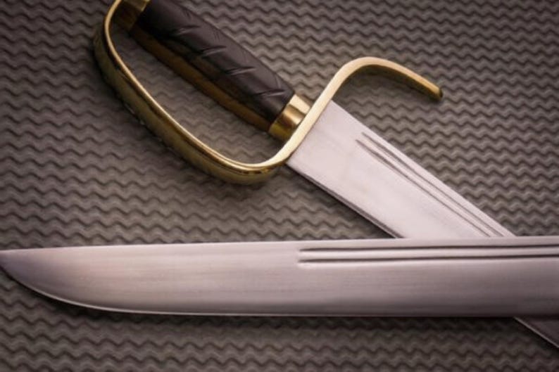 K&ouml;nnte beinhalten: Nahaufnahme eines S&auml;bels mit einem gebogenen, goldfarbenen Handschutz und einem schwarzen Griff. Die Klinge ist silberfarben und hat eine dekorative Rille. Das S&auml;bel liegt auf einer strukturierten, grauen Oberfl&auml;che.