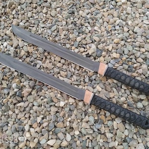 May include: Two grey swords with black wrapped handles and copper-coloured guards are displayed on a bed of small, multicoloured stones. The blades are rectangular with a sharp edge. The handles are textured for grip.