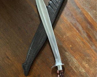 May include: A large, curved blade knife with a silver blade and a wooden handle. The knife is paired with a black sheath. The handle is intricately carved. The knife and sheath are resting on a wooden surface.