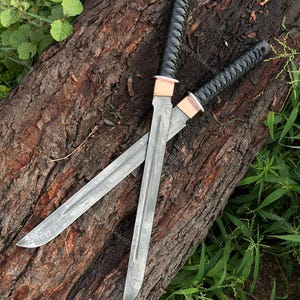 May include: Two straight-edged swords with silver-toned blades featuring a visible folded metal pattern. Each sword has a black cord-wrapped handle, a copper-coloured collar, and a pointed tip. The blades are positioned in an overlapping cross shape.