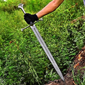 Puede incluir: Una espada larga y plateada con una hoja estampada y un mango adornado se sostiene contra un fondo de follaje verde. El mango de la espada presenta una guarda cruzada y un pomo decorativo. La hoja tiene un patrón texturizado detallado.
