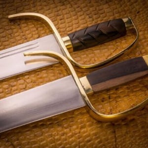 K&ouml;nnte beinhalten: Zwei Wing Chun Schwerter mit polierten Stahlklingen und messingfarbenen Griffen. Die Griffe sind aus dunkelbraunem Holz. Die Schwerter werden auf einer strukturierten, braunen Oberfl&auml;che pr&auml;sentiert.