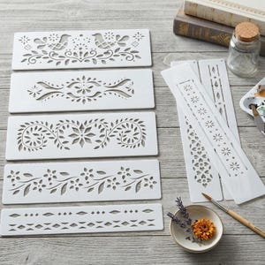 May include: A set of seven white rectangular plastic stencils featuring various laser-cut patterns including birds, floral vines, geometric shapes, and repeating leaf motifs. The stencils are arranged in a stack and side-by-side layout.